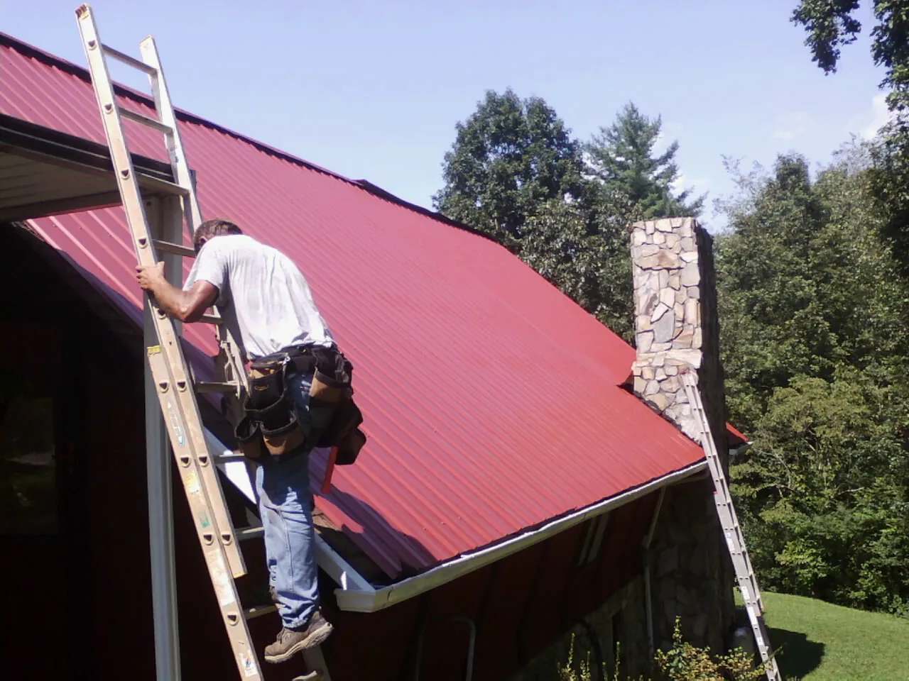 Roofer on a ladder beside a red metal roof and stone chimney — Soffit & Fascia Repair by Roofing Brothers in Farmington